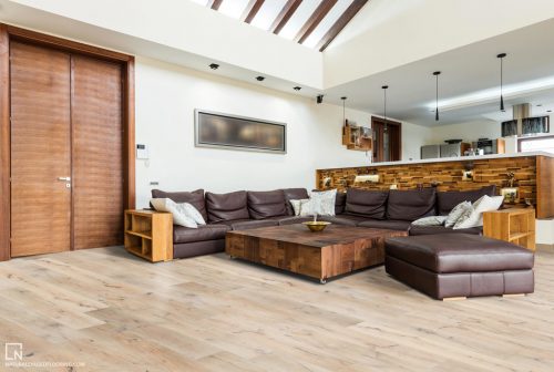 hardwood floor in living room with brown leather couch and ottoman, large wood block table, wood accents and kitchen in the background