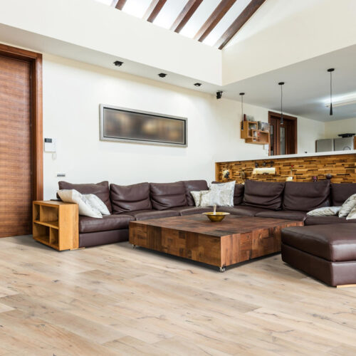 hardwood floor in living room with brown leather couch and ottoman, large wood block table, wood accents and kitchen in the background
